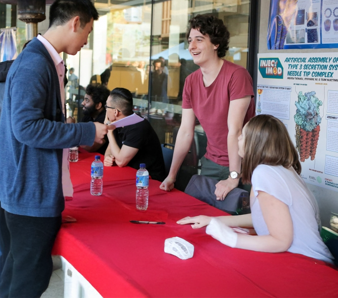 A group of people gathered around a table with red tablecloths, engaged in conversation. Some are standing while others are seated. Behind them is a large poster titled ‘Artificial Assembly of the Type 3 Secretion System Needle Tip Complex,’ featuring scientific diagrams. Bottles of water are on the table.